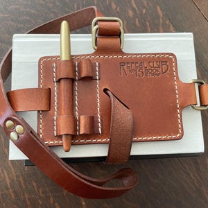 May include: A brown leather book strap with a pen holder and a brass buckle. The strap is labeled "Repeal Club No. 15 Book Strap".