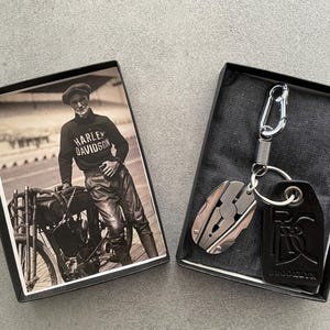 Op de afbeelding: Een zwarte leren sleutelhanger met een zilveren multitool en een Harley Davidson logo. De sleutelhanger zit in een zwarte doos met een foto van een man die een Harley Davidson shirt draagt en een motorfiets rijdt.