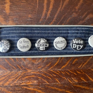 May include: A set of six vintage black and white button pins in a box. The pins feature slogans related to the prohibition era, including "Real Club 18", "No Beer Norwalk", "Bring Back Beer", "I Am Against Prohibition", "Vote Dry", and "We Want Beer".