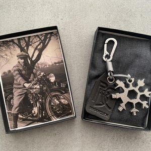 Pode incluir: Uma caixa preta com uma foto vintage de um homem a conduzir uma motocicleta. A caixa está aberta e mostra um chaveiro multiferramentas em forma de floco de neve prateado com uma etiqueta de couro preto que diz "PRODESIGN".
