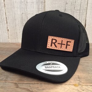 May include: Black and brown baseball cap with a leather patch that says "R+F" and a silver sticker that says "The Classics Snapback Yupoong".