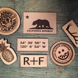 May include: A collection of seven leather patches with various designs. The patches feature a skull with a flower crown, a bear, a set of antlers, a pineapple, a smiley face with a lemon slice, coordinates, and the letters "R+F".