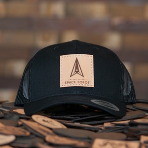 Könnte beinhalten: Schwarze Mesh-Trucker-Kappe mit einem braunen Leder-Patch mit dem Logo der US Space Force. Das Logo ist ein Delta mit einem Stern und dem Text "UNITED STATES SPACE FORCE".