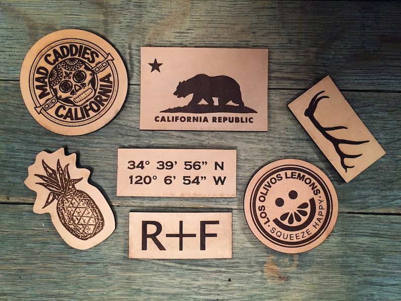 May include: Seven leather patches with various designs. One patch features a skull with the text "Mad Caddies Old California". Another patch features a bear with the text "California Republic". A third patch features a pineapple. A fourth patch features the text "34&deg; 39' 56" N 120&deg; 6' 54" W". A fifth patch features the text "R+F". A sixth patch features a deer antler. A seventh patch features a lemon with the text "Solivios Lemons, Squeeze Happy."