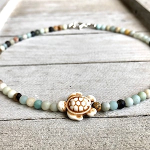 May include: A light blue and brown beaded necklace with a white turtle charm. The necklace is made of natural stones and is perfect for a beachy look.