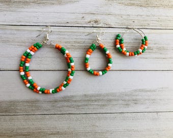 May include: Three pieces of jewelry featuring orange, white, and green seed beads arranged in a repeating pattern. The set includes one large circular hoop earring, one smaller teardrop-shaped hoop earring, and one small circular hoop earring.