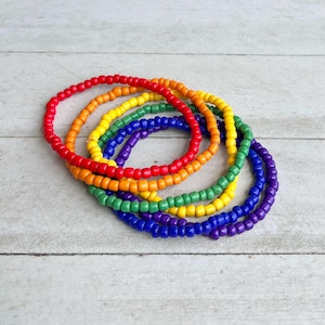 May include: A set of five circular bracelets made from small, smooth, round beads in vibrant colors. The bracelets feature individual strands of red, orange, yellow, green, blue, and purple beads, arranged in a stacked, overlapping circular formation.
