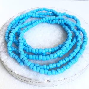 May include: Several strands of light blue beads are arranged in a circular pattern on a white and gray surface. The beads are small, uniform in size, and have a slightly matte finish. The overall aesthetic is simple and clean.
