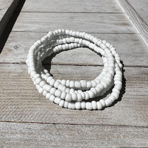 May include: A collection of white beaded bracelets, coiled on a weathered wooden surface. The beads are small and uniform, creating a smooth, continuous texture. The bracelets are a simple, elegant accessory.