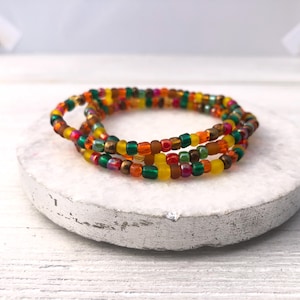 May include: A multi-strand bracelet composed of small, round glass seed beads in a vibrant palette of yellow, orange, red, green, and brown. The beads are arranged in a repeating pattern, creating a textured, circular band that sits in a coiled loop.