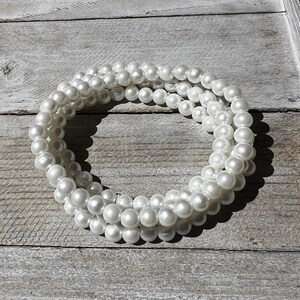 May include: A multi-strand bracelet composed of numerous small, round, white faux pearls. The beads are uniform in size and arranged in a circular, layered formation, creating a continuous loop of reflective, smooth, white spherical elements.