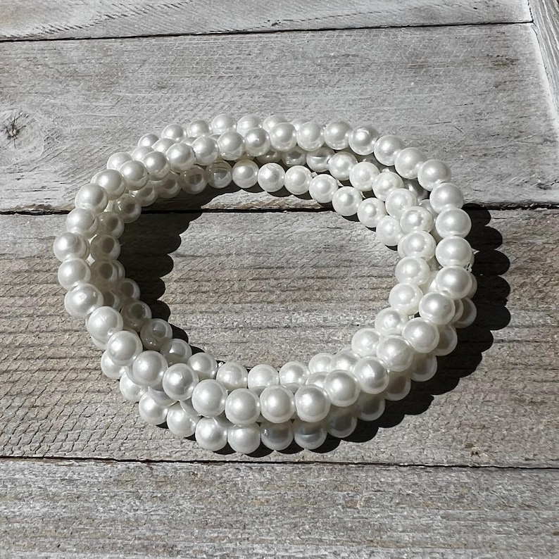May include: A multi-strand bracelet composed of numerous small, round, white faux pearls. The beads are uniform in size and arranged in a circular, layered formation, creating a continuous loop of reflective, smooth, white spherical elements.