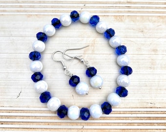 May include: A matching jewelry set featuring a bracelet and earrings. The bracelet and earrings are made with white and dark blue beads. The earrings have silver-colored hooks. The jewelry set is displayed on a light-colored wooden surface.