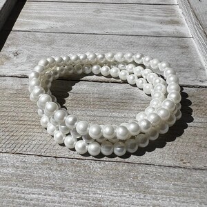May include: A multi-strand bracelet composed of three rows of smooth, round, white faux pearls. The beads are uniform in size and have a lustrous, reflective finish, creating a continuous circular shape that is flexible and lightweight in appearance.