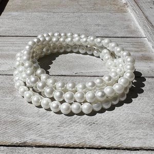 May include: A multi-strand bracelet composed of numerous smooth, round, white faux pearls. The pearls are uniform in size and arranged in a circular, layered formation, creating a continuous loop of reflective, glossy white beads.