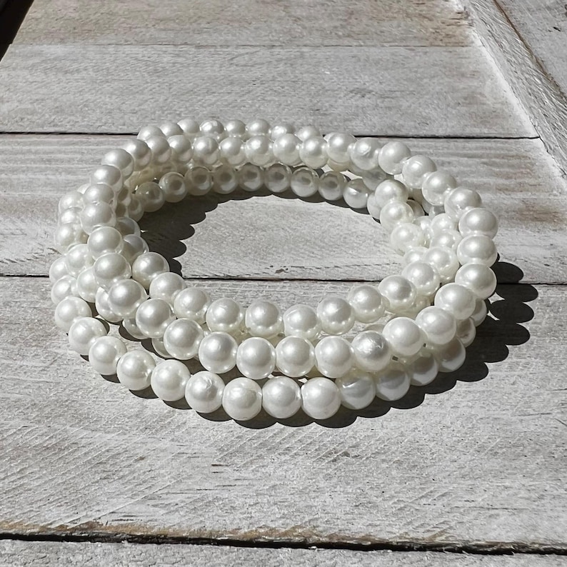 May include: A multi-strand bracelet composed of numerous smooth, round, white faux pearls. The pearls are uniform in size and arranged in a circular, layered formation, creating a continuous loop of reflective, glossy white beads.