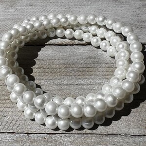 May include: A multi-strand bracelet composed of numerous round, white, lustrous faux pearls. The beads are uniform in size and arranged in a circular, layered formation, creating a continuous loop of reflective, smooth, spherical white pearl-like elements.