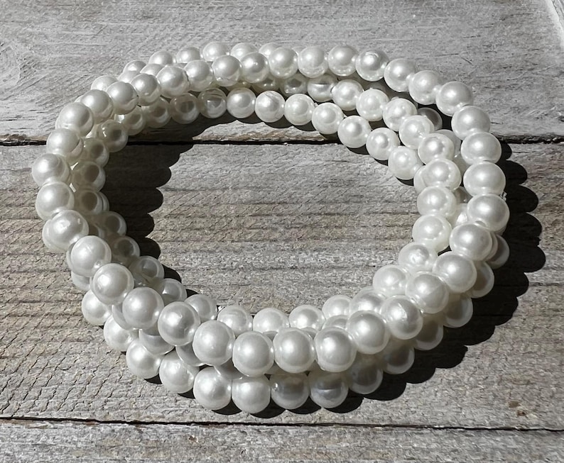 May include: A multi-strand bracelet composed of numerous round, white, lustrous faux pearls. The beads are uniform in size and arranged in a circular, layered formation, creating a continuous loop of reflective, smooth, spherical white pearl-like elements.