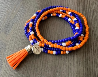 May include: A multi-strand beaded bracelet featuring vibrant orange, royal blue, and white seed beads. The bracelet includes a silver-toned circular charm engraved with the letter N and a matching orange suede-like tassel attached to the clasp.