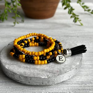 May include: A multi-strand bracelet made of small, round, yellow and black beads. The bracelet features a silver-toned circular charm with the letter E printed in black, and a black suede tassel attached to a silver-toned metal cap.