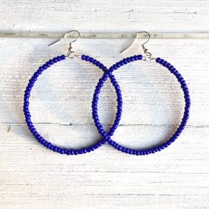 May include: A pair of hoop earrings featuring small, vibrant royal blue seed beads strung along a circular metal frame. Each earring is attached to a simple silver-toned metal fishhook ear wire, designed for hanging from the earlobe.