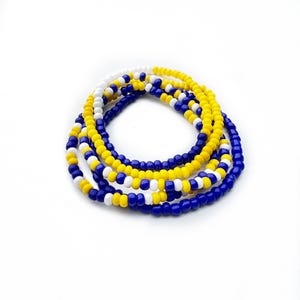 May include: A set of three circular beaded bracelets featuring small, round, opaque beads in solid colors of bright yellow, royal blue, and white. The beads are strung together in a repeating pattern, creating a layered, flexible, and lightweight accessory.