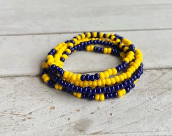 May include: A circular bracelet composed of small, round beads in alternating segments of bright yellow and deep navy blue. The beads are strung together in a continuous loop, creating a vibrant, multi-colored pattern that encircles the entire piece.