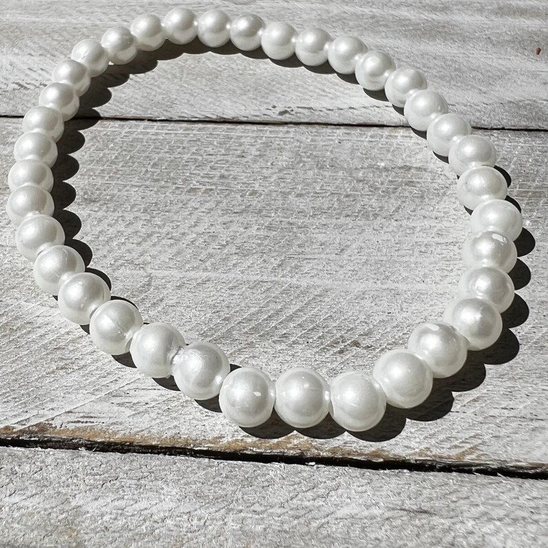 May include: A circular bracelet composed of numerous smooth, round, white faux pearls of uniform size. The beads are strung together in a continuous loop, reflecting light on their glossy surfaces, creating a simple and classic jewelry accessory.