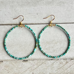 May include: A pair of hoop earrings featuring small, round, faceted turquoise-colored beads strung along a thin gold-toned wire frame. Each earring is suspended from a simple gold-toned metal hook, creating a circular design with a vibrant blue-green color palette and a textured, beaded appearance.