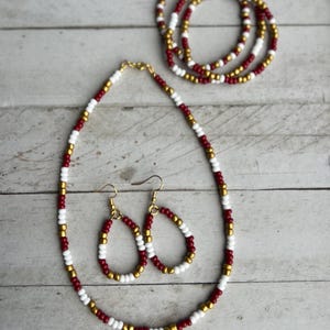 May include: A jewelry set consisting of a beaded necklace, a pair of teardrop-shaped beaded earrings, and a beaded bracelet. Each piece is crafted from small, round beads in alternating colors of deep red, crisp white, and metallic gold.