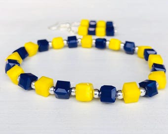 May include: A bracelet and earrings set featuring yellow and navy blue cube-shaped beads. The bracelet is strung with silver-colored beads. The earrings have a similar design. The jewelry set is on a white surface.