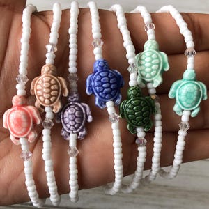 May include: Six beaded bracelets featuring white seed beads and clear crystal accents. Each bracelet includes a single ceramic sea turtle charm in a different color: coral, tan, purple, blue, dark green, light green, and mint green.