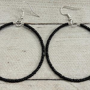 May include: A pair of hoop earrings featuring a circular frame covered in small, uniform black beads. Each earring is attached to a silver-toned metal fishhook style ear wire, allowing the circular beaded hoops to hang vertically from the ear.