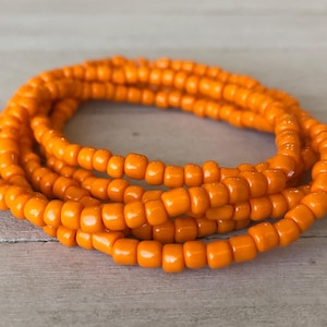 May include: A coiled bracelet made of small, uniform, opaque orange seed beads. The beads are strung together in a continuous loop, creating a vibrant, solid-colored circular accessory with a smooth, glossy finish throughout the entire piece.
