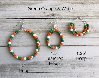 May include: Three pairs of earrings featuring green, orange, and white seed beads. The set includes a 2-inch hoop, a 1.5-inch teardrop hoop, and a 1.25-inch hoop. Each earring is constructed with silver-toned metal hooks and wire frames.