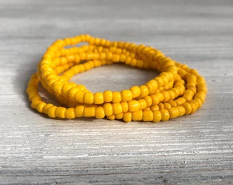 May include: A stack of multiple bracelets made from small, round, vibrant yellow beads. The beads are uniform in size and shape, creating a continuous circular pattern. The bracelets are arranged in a loose, overlapping coil, showcasing the bright, solid yellow color of the individual glass or plastic beads throughout the entire accessory set.