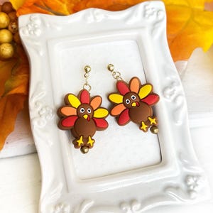 May include: A pair of dangle earrings featuring brown turkey charms with multi-colored feathers in red, orange, and yellow. The turkeys have white eyes, yellow beaks, and yellow feet. Each earring hangs from a gold-toned metal post and hook.