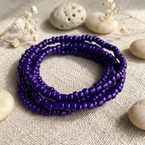 May include: A coiled bracelet made of small, uniform, deep purple glass beads. The circular strand is composed of multiple loops of these smooth, opaque beads, creating a continuous, vibrant purple ring that stands out against the neutral background.