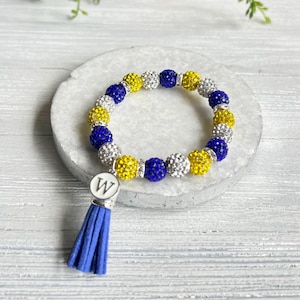 May include: A stretch bracelet featuring alternating round beads in blue, yellow, and silver, all covered in small sparkling crystals. A silver-toned circular charm with the letter W engraved in black hangs from a blue suede-like fabric tassel.