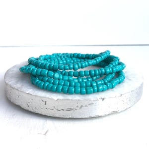 May include: A coiled strand of small, round, opaque turquoise beads. The beads are uniform in size and shape, strung together to form a continuous loop. The vibrant blue-green color of the beads is consistent throughout the entire strand.