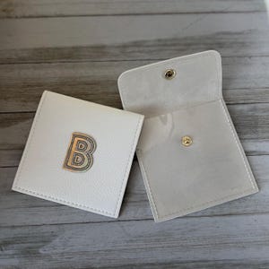 May include: Two square white pouches with a textured finish. One pouch is closed and features a prominent gold-toned letter B emblem on the front. The other pouch is open, revealing a smooth interior and a single gold-toned metal snap button closure.