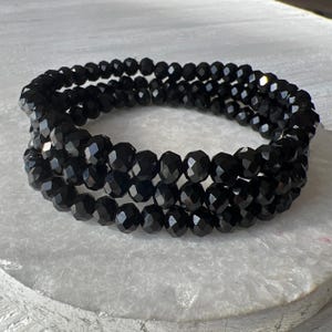 May include: A multi-strand bracelet composed of small, round, faceted black beads. The beads have a reflective, glossy finish and are arranged in a circular, layered formation, creating a continuous, uniform appearance throughout the entire piece.