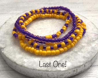 May include: A stack of beaded bracelets featuring alternating strands of small, round, opaque yellow beads and translucent purple beads. The bracelets are arranged in a circular pattern, and the text "Last One!" is printed below the jewelry.