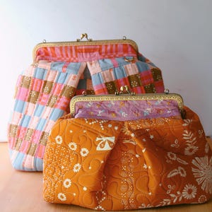 Puede incluir: Dos bolsos de tela acolchados con cierres de latón. El bolso más grande es azul, rosa y marrón con un patrón de cuadros. El bolso más pequeño es naranja con un patrón floral.