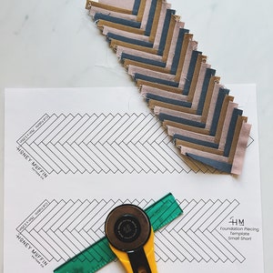May include: A foundation piecing template for a quilt pattern called "Honey Muffin" with a rotary cutter, ruler, and fabric squares in shades of brown, gray, and pink.