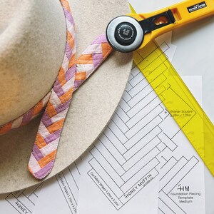 May include: A close-up of a quilting project with a yellow ruler, a rotary cutter, and a white hat with a pink and orange braided trim. The project includes a template for a foundation piecing quilt pattern called "Honey Muffin" with instructions to cut fabric strips 1.25 inches by 3.25 inches.