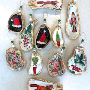 May include: A collection of handcrafted Christmas ornaments made from oyster shells. Each shell features a unique design, including a fox, Santa hat, Christmas tree, and floral patterns. The shells have gold-painted edges and are strung with beads.