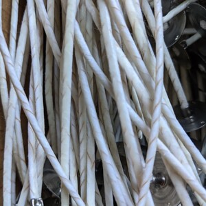 May include: Close-up of a bundle of white candle wicks with a thin, twisted design and a brown thread running through the center. The wicks are attached to small, silver metal tabs.