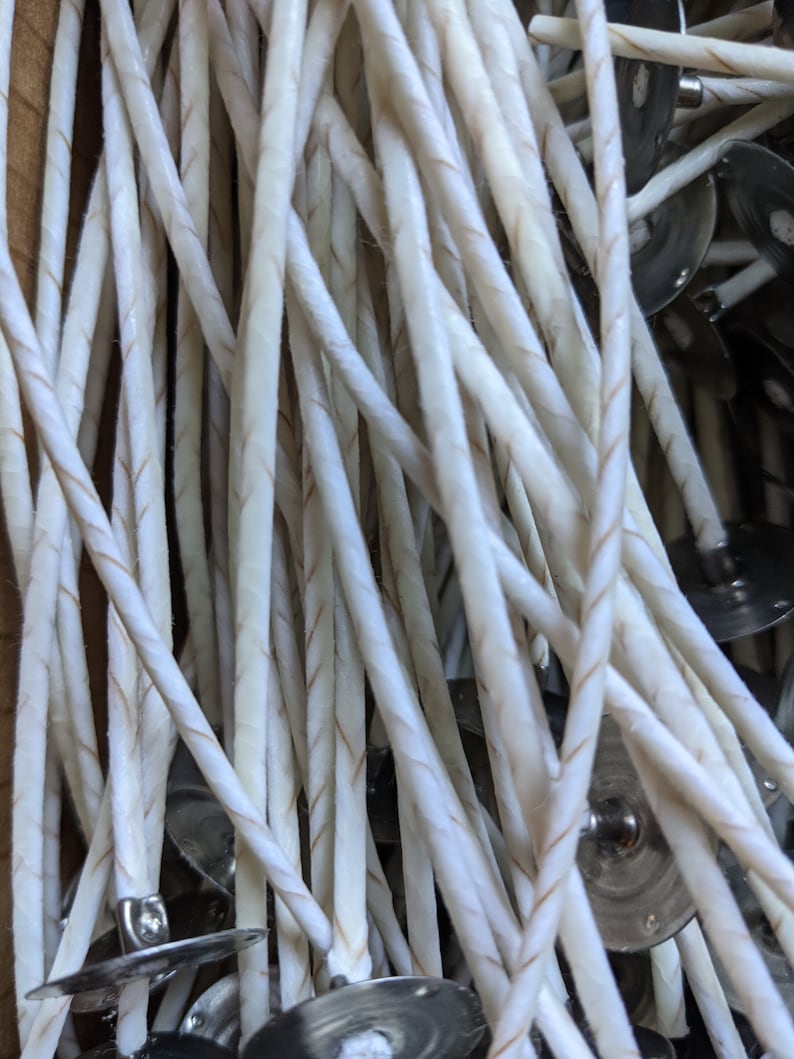 May include: Close-up of a bundle of white candle wicks with a thin, twisted design and a brown thread running through the center. The wicks are attached to small, silver metal tabs.