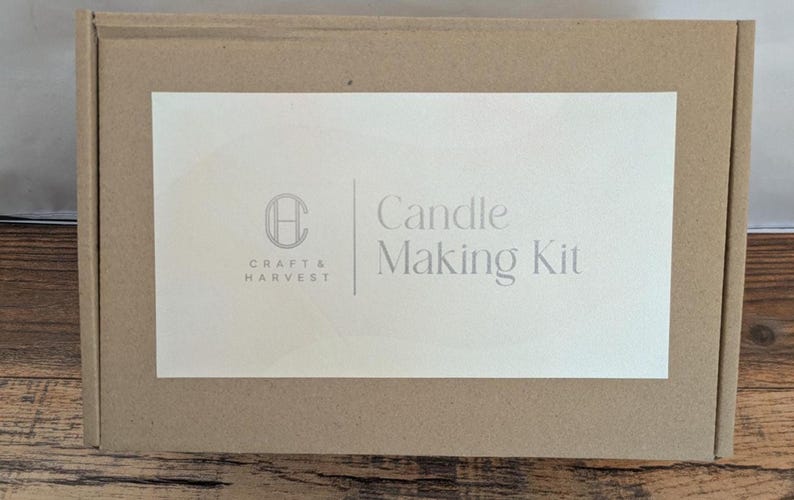 Pu&ograve; includere: Una scatola rettangolare di cartone marrone con un'etichetta bianca. L'etichetta presenta il testo "Candle Making Kit" e il logo "Craft & Harvest". La scatola &egrave; su una superficie di legno.